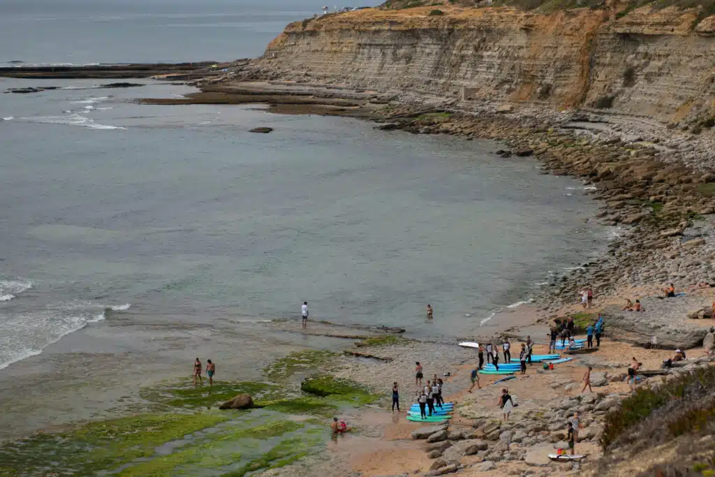 People gather on a rocky beach—one of the Best Beaches in Ericeira—with blue surfboards near coastal cliffs. Some enter the water, others stand onshore amid tide pools and seaweed, as the ocean extends into the distance.