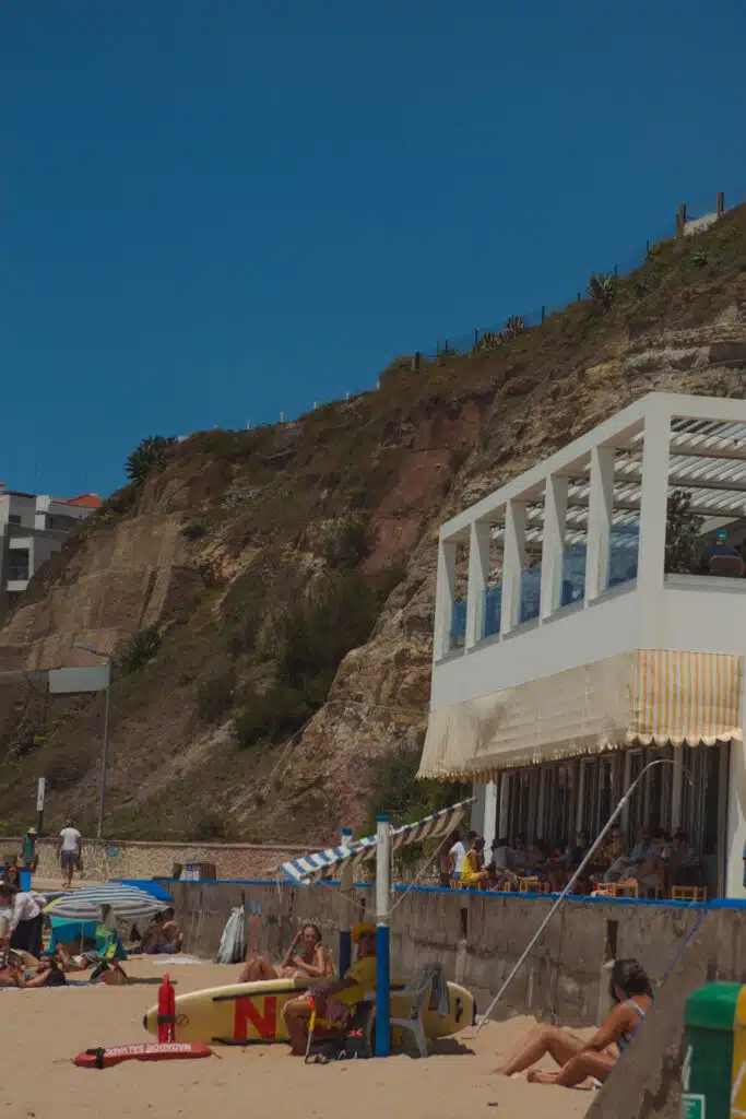 People relax on a sandy beach near a white seaside building with a striped yellow awning, one of the Best Beaches in Ericeira. Behind them, a rocky, green hillside rises under a clear blue sky. Beach umbrellas and a lifeguard board are visible.