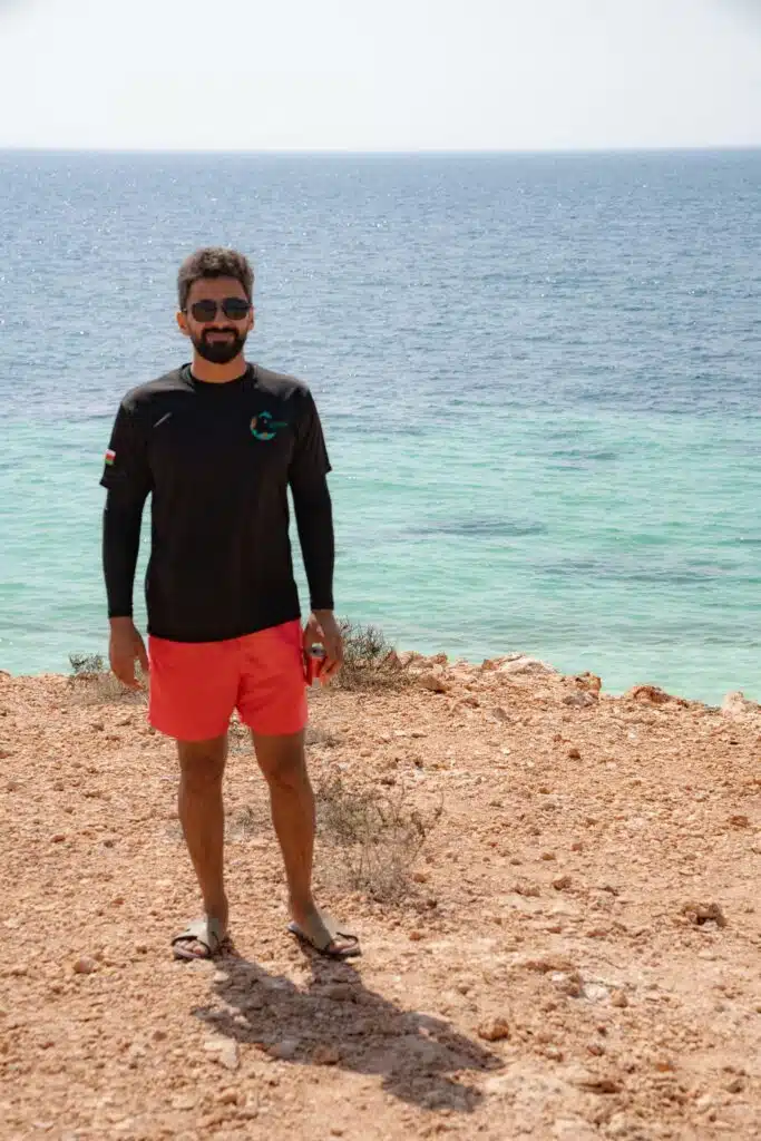 A man wearing sunglasses, a black long-sleeve shirt, and red shorts stands on the rocky beach of the Daymaniyat Islands with turquoise sea water and a clear sky in the background.