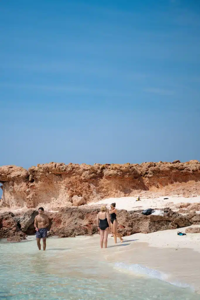 Three people in swimsuits stand and walk along a sandy beach with clear blue water on the Daymaniyat Islands, rocky terrain, and a bright blue sky in the background. Towels and items are placed on the rocks behind them.