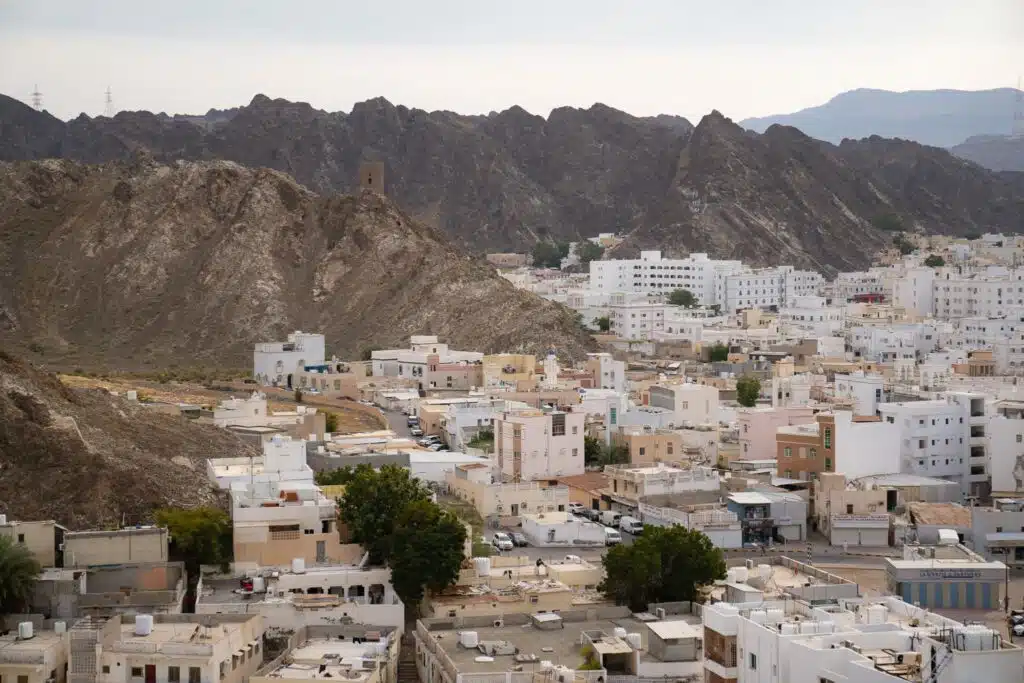 A view of white, flat-roofed buildings clustered at the base of rocky mountains, with a historic watchtower on a hill and cloudy sky above in Muscat, Oman, gateway to the stunning Daymaniyat Islands.