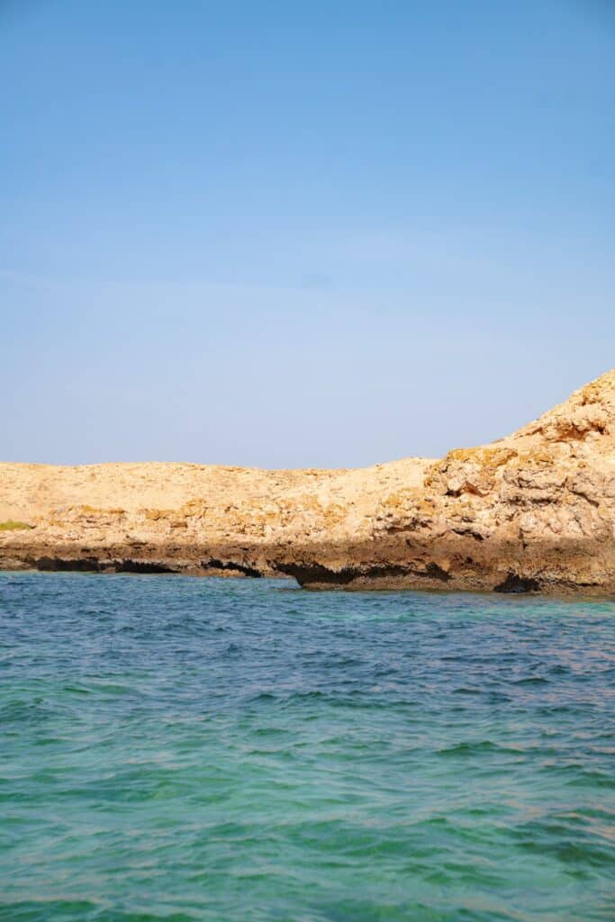 Clear blue-green water in the foreground frames the rocky, beige coastline of the Daymaniyat Islands, with a bright blue sky in the background.