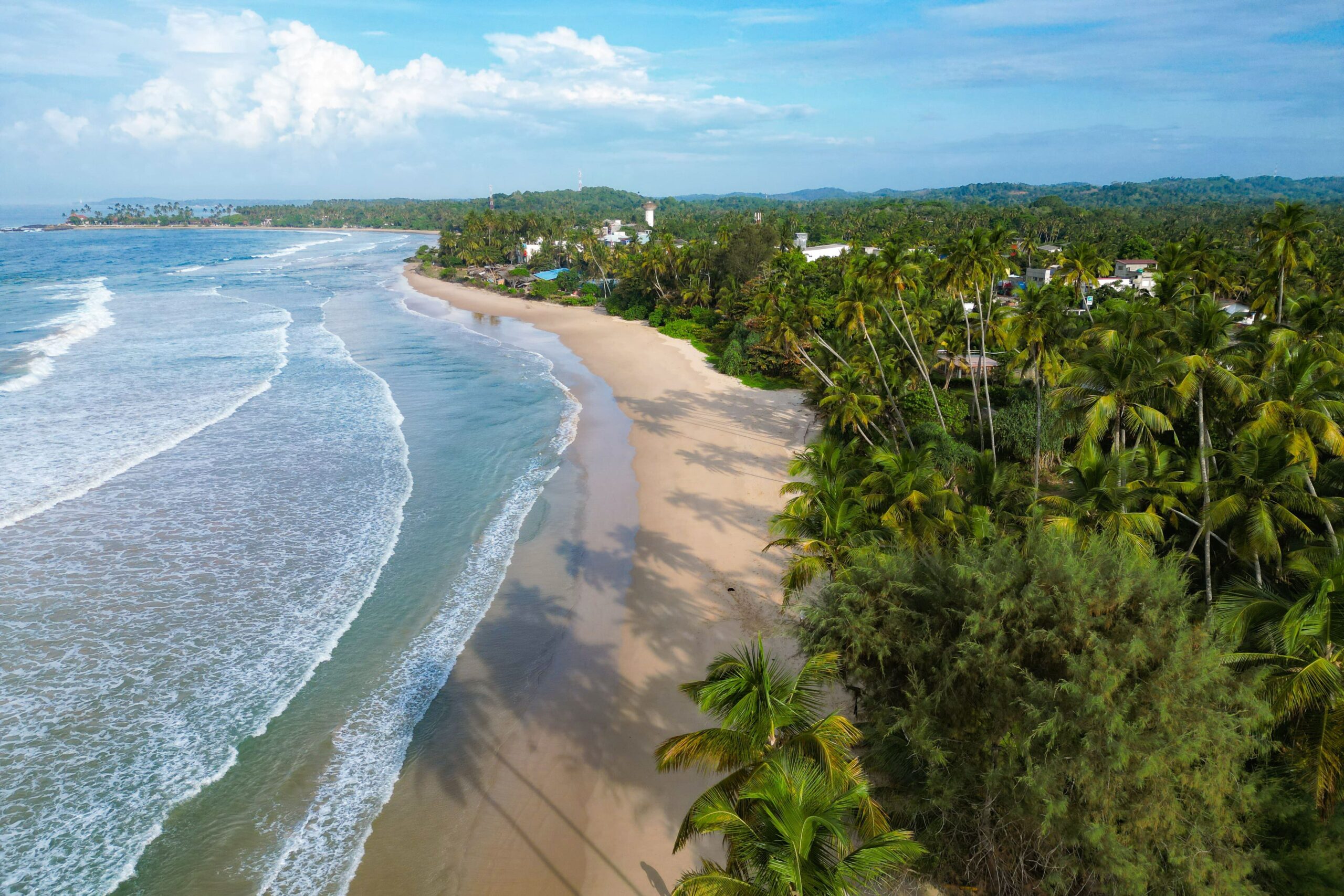 Aerial view of a tropical beach with gentle waves, golden sand, and lush green palm trees. Discover houses and greenery along the coastline under a partly cloudy blue sky—perfect for exploring the best Things To Do In Hiriketiya.