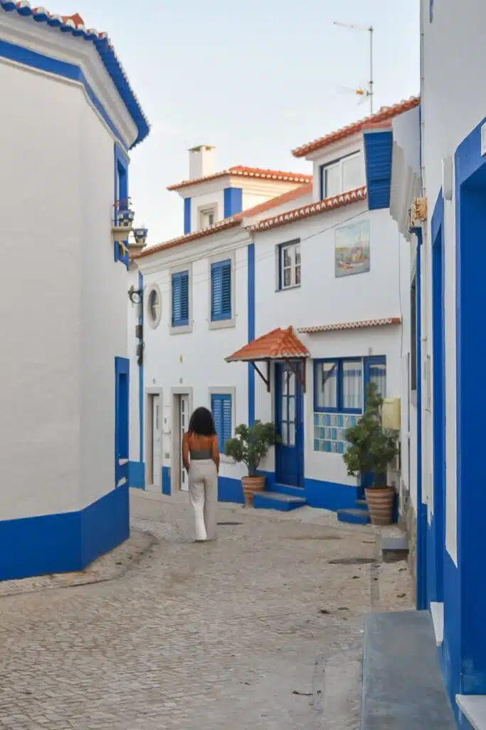 A digital nomad in Ericeira with long hair walks down a narrow cobblestone street lined with white buildings featuring blue trim and red-tiled roofs on a clear day.