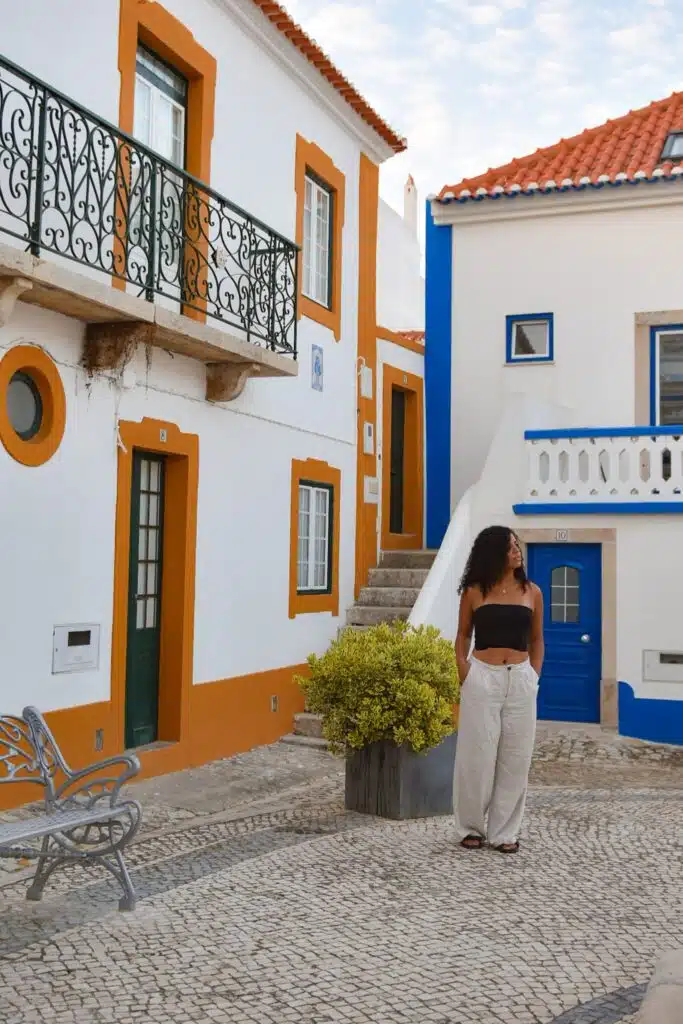 A Digital Nomad in Ericeira, wearing a black top and light pants, stands on a cobblestone street before colorful buildings with orange trim, green doors, and a blue railing under a partly cloudy sky.