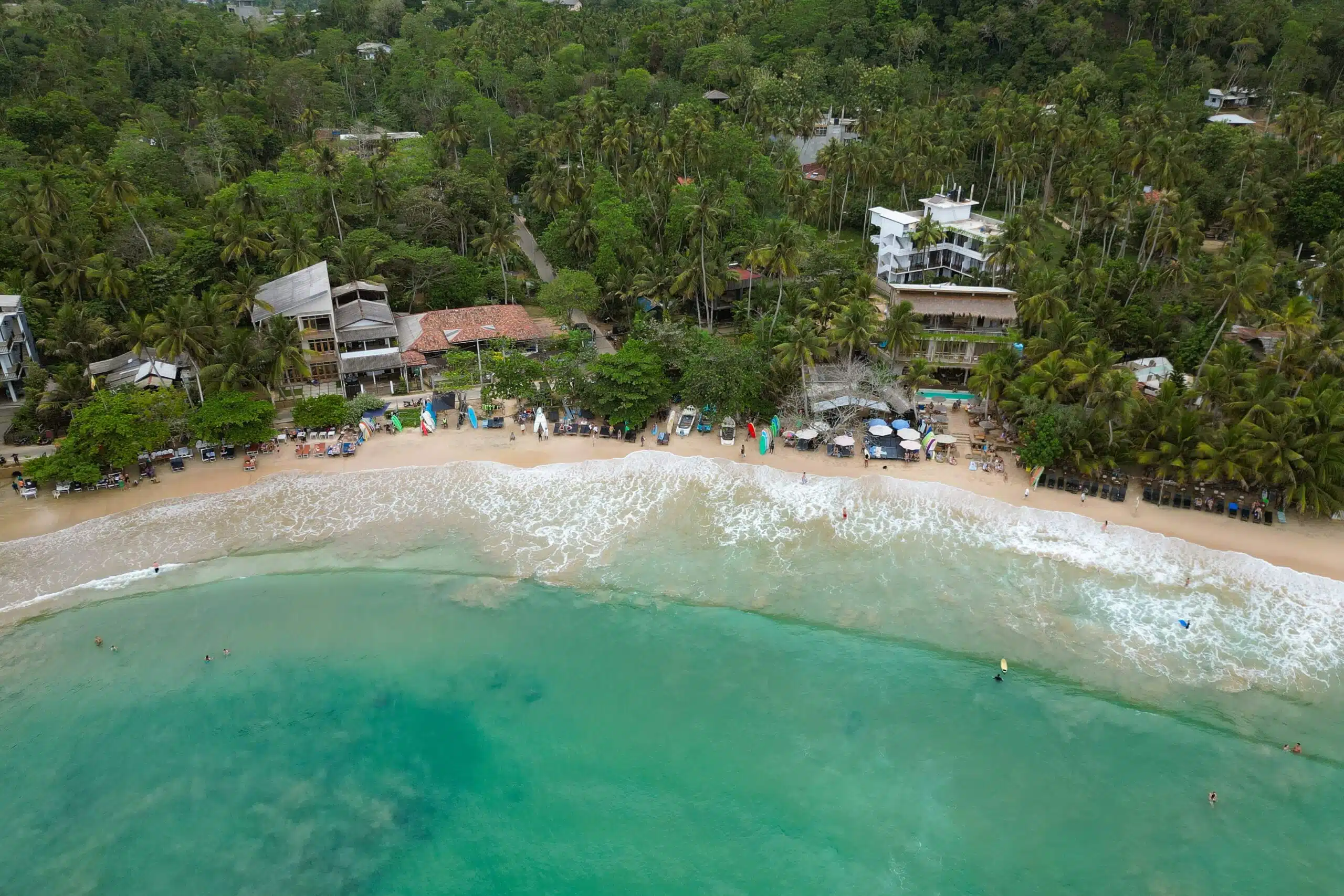 Aerial view of a tropical beach with turquoise water, gentle waves, and people swimming. Beachfront buildings and palm trees line the shore—one of the top things to do in Hiriketiya for nature lovers, with lush forest stretching into the background.
