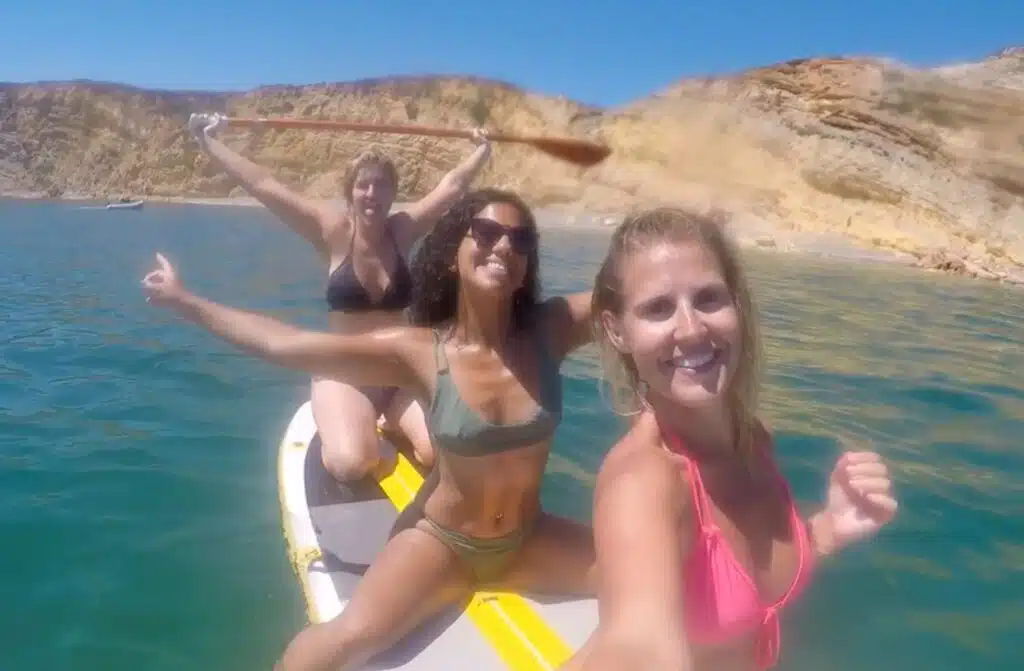 Three women in swimsuits pose and smile on a paddleboard during a Boat Tour in Lagos, floating on clear blue water with rocky cliffs and a sunny sky behind them. One woman celebrates by holding a paddle above her head.