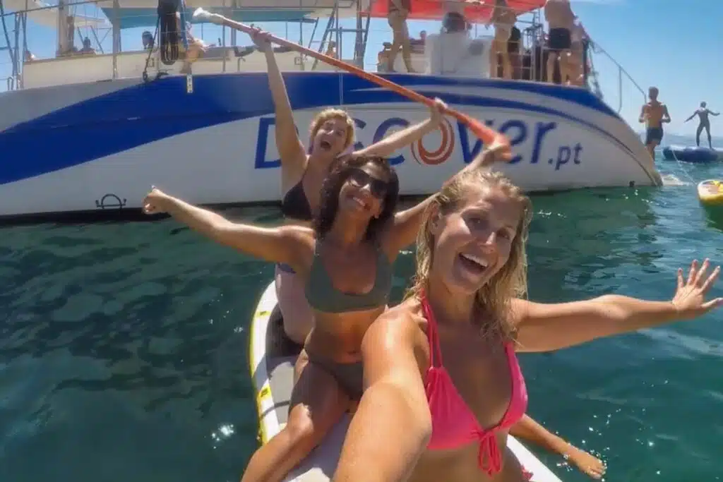 Three women in swimsuits smile and pose for a selfie on a paddleboard in the ocean during a Boat Tour in Lagos, with a large boat and several others enjoying the sunny day in the background.
