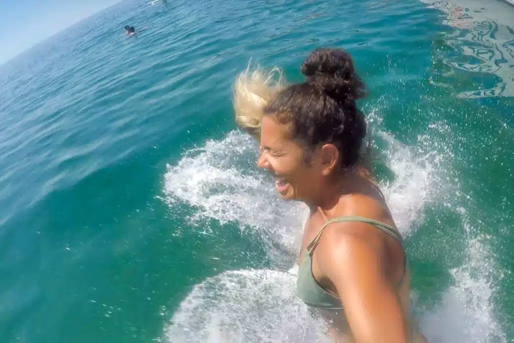 Two women in swimsuits laugh as they jump into the ocean during a Boat Tour in Lagos, splashing water around them. The sun shines on the clear blue-green water, with another person swimming in the distance.