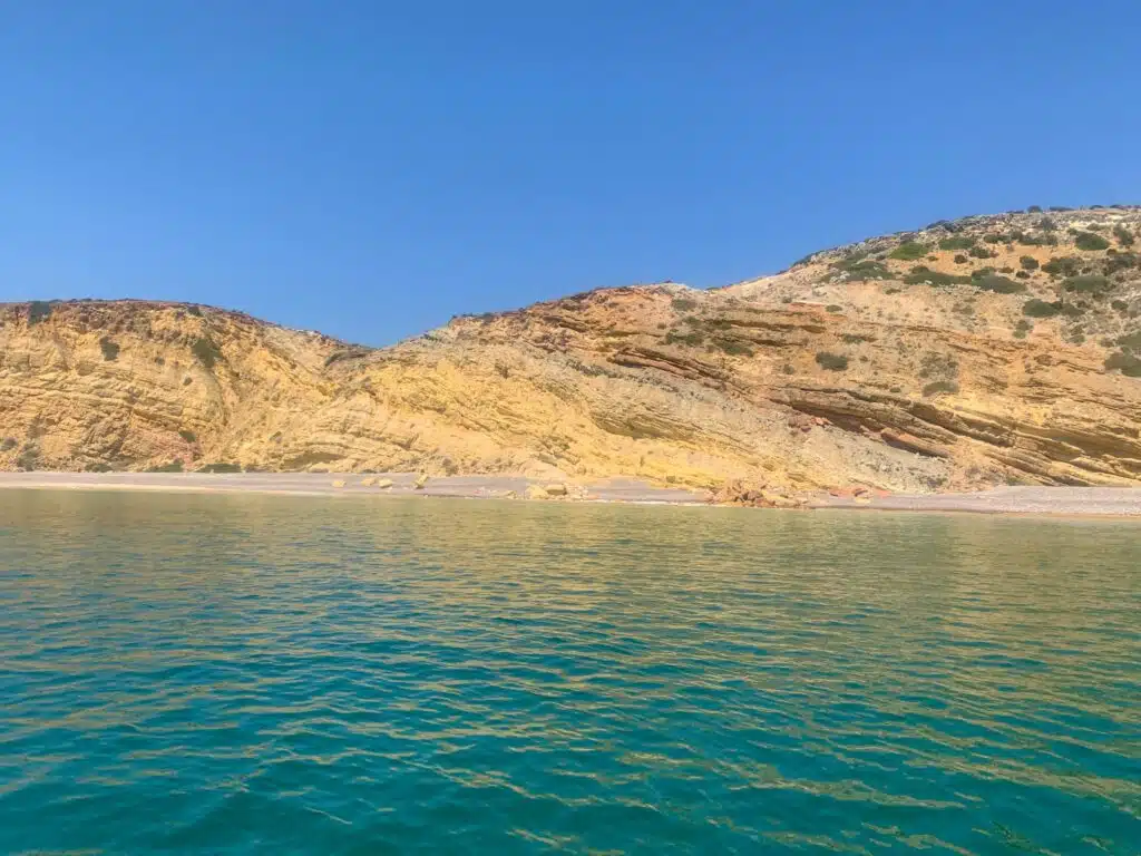 Rocky, multicolored cliffs with layered patterns rise above a pebble beach, meeting clear turquoise water under a bright blue sky—perfect scenery to admire during a boat tour in Lagos. Sparse vegetation dots the hillside.