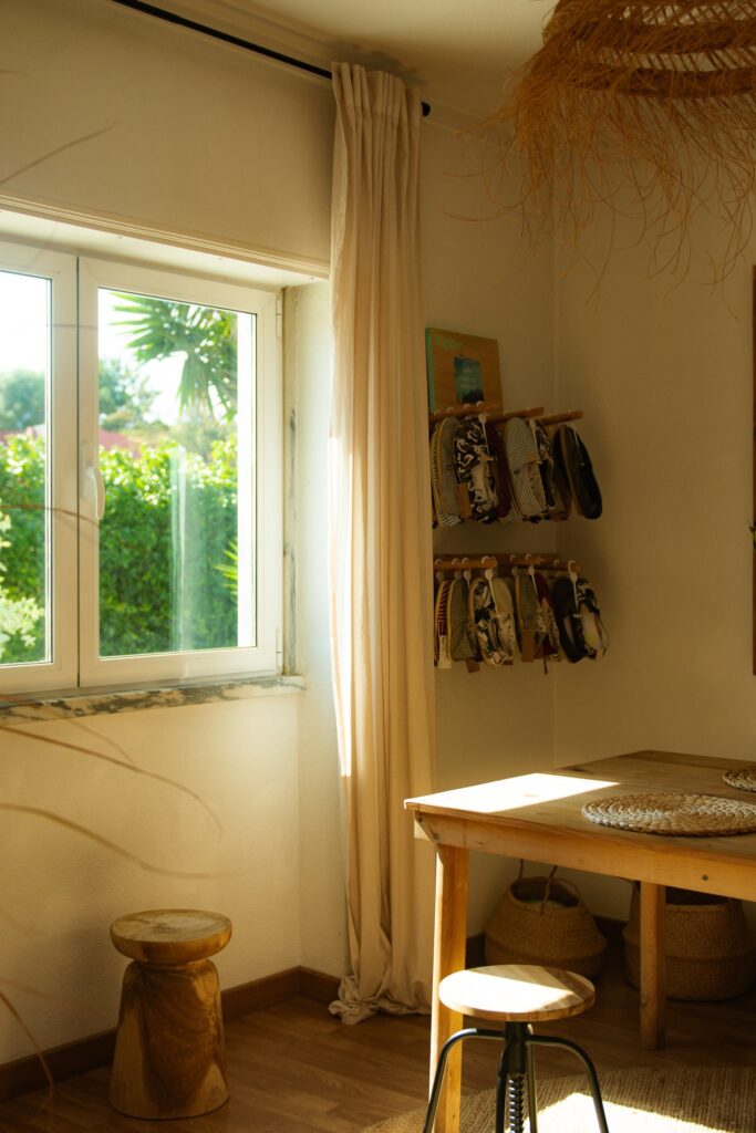Bright, sunlit room with a wooden table and stool, cream curtains by the windowโa cozy retreat for a digital nomad in Ericeira. Small bags or pouches hang on the wall; baskets are placed neatly under the table.