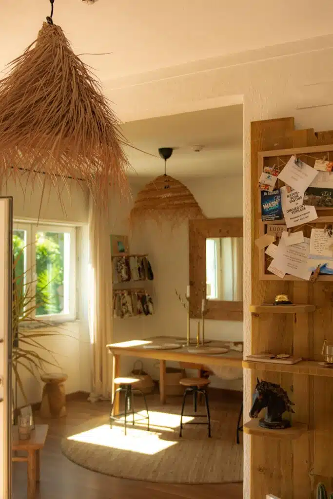 Bright, cozy room with wooden furniture, woven light fixtures, a round rug, and sunlit window—perfect for a Digital Nomad in Ericeira. A bulletin board with notes hangs on the right, and a desk with stools sits in the background.