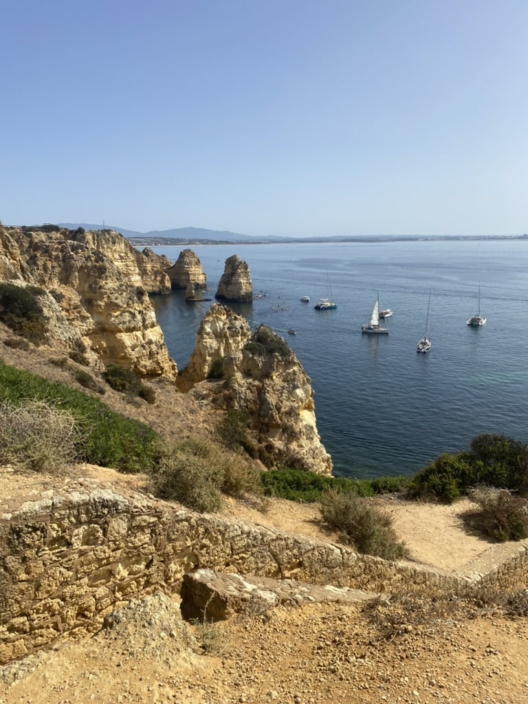 Rocky cliffs overlook calm blue water where several sailboats are anchored near tall sea stacks—a perfect scene for a Boat Tour in Lagos. Green shrubs line the rocky shore beneath a clear, sunny sky.