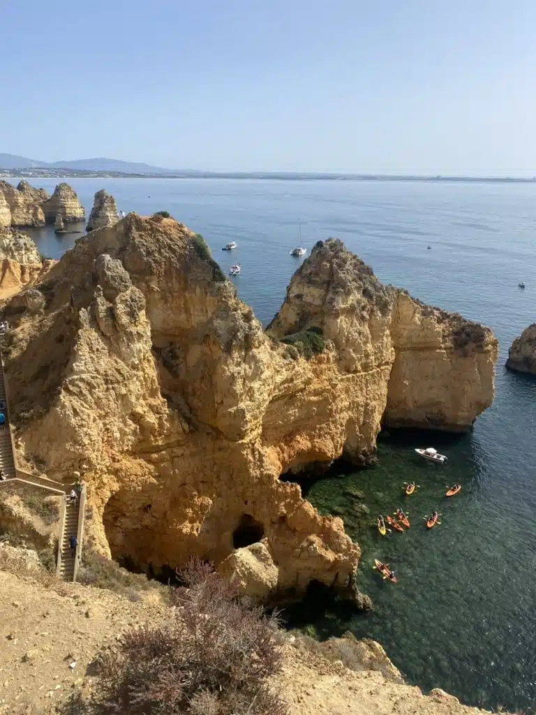Aerial view of rocky cliffs and clear blue water with several kayakers paddling near the base, a few boats in the distance, and a Boat Tour in Lagos under a clear sky.