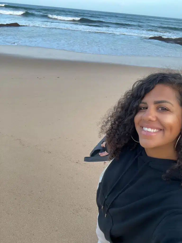 A woman with curly hair smiles while taking a selfie on a sandy beach in Ericeira, waves and rocks behind her. Sporting a black and white hoodie and large hoop earrings, she captures the digital nomad lifestyle by the sea.