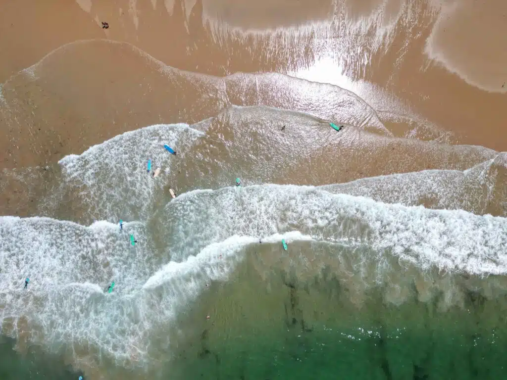 Aerial view of surfers with boards in the shallow waves near a sandy beach—one of the Best Beaches in Ericeira—where white foam and green water shimmer under sunlight reflecting off the wet sand.