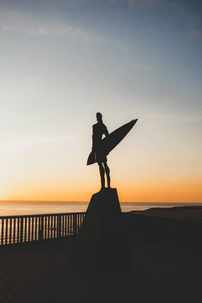 Silhouette of a surfer statue holding a surfboard, standing on a pedestal against a sunset sky near the ocean—an iconic scene near the Best Beaches in Ericeira, with calm water and a railing in the background.