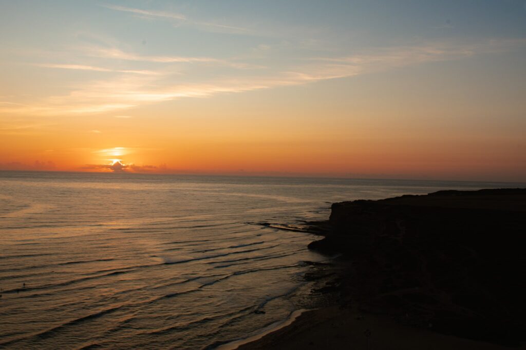 A serene sunset over the ocean, with gentle waves approaching a dark, rocky shoreline on the right and a colorful sky blending orange and blue huesโcapturing the tranquil beauty of the Best Beaches in Ericeira.