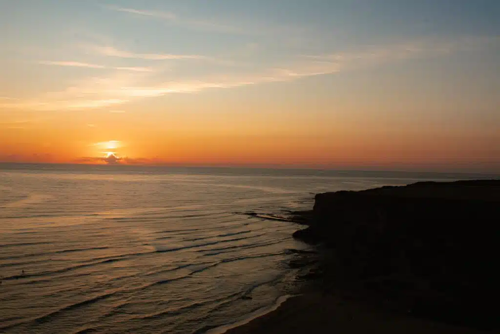 A serene sunset over the ocean, with gentle waves approaching a dark, rocky shoreline on the right and a colorful sky blending orange and blue hues—capturing the tranquil beauty of the Best Beaches in Ericeira.