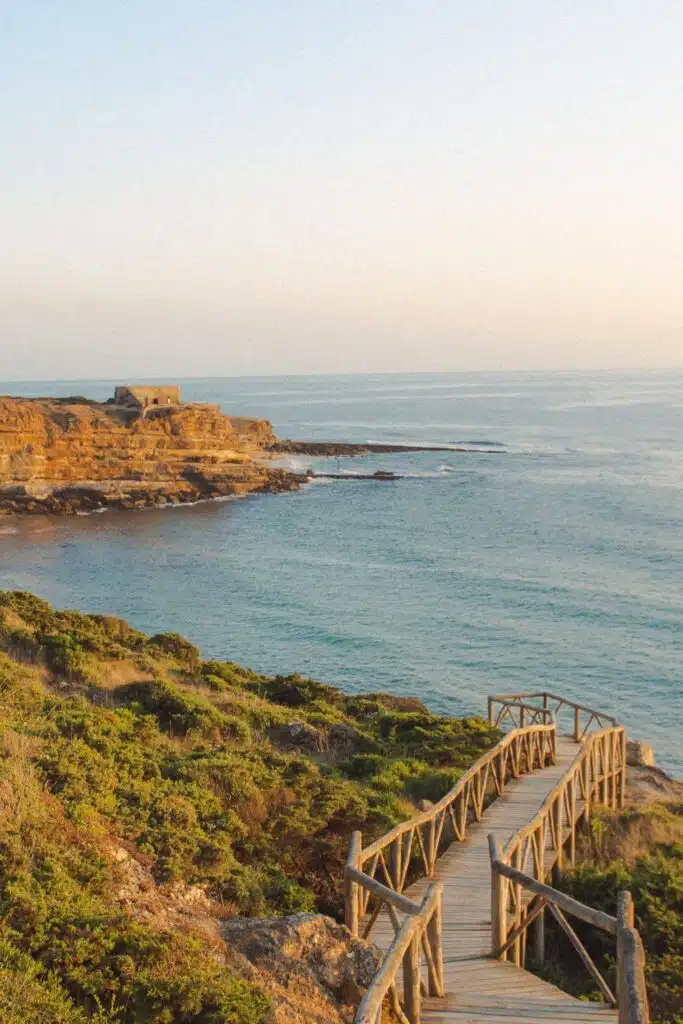 A wooden boardwalk with railings leads down a grassy, green hillside toward a rocky coastline and calm blue sea under a clear sky at sunset—classic scenery near some of the Best Beaches in Ericeira. A small stone building sits on the distant cliff.