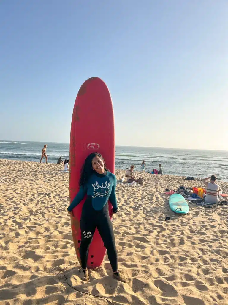 A woman in a wetsuit smiles while standing in front of a tall red surfboard on the sandy shores of one of the Best Beaches in Ericeira, with the ocean and people relaxing or preparing to surf under a clear sky in the background.