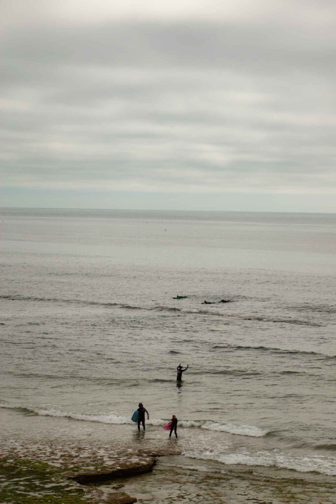 Two people, one adult and one child, stand near the shore with surfboards on one of the Best Beaches in Ericeira, while three more surfers are farther out in the gray, calm ocean under a cloudy sky.