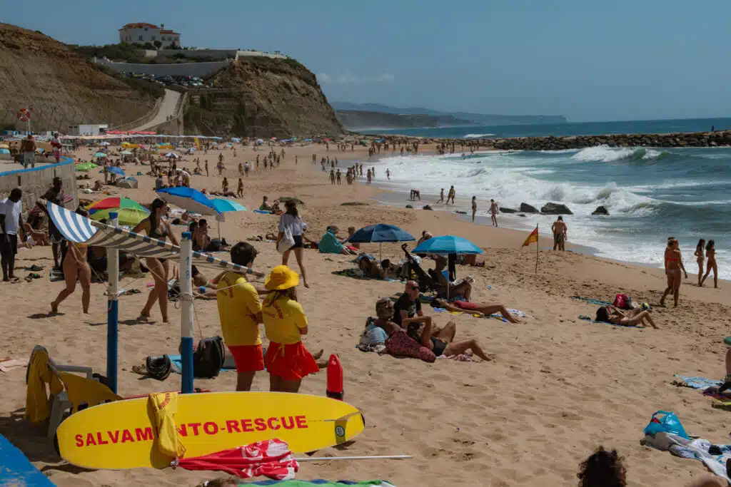 Crowded beach scene with people sunbathing and relaxing under umbrellas at one of the Best Beaches in Ericeira. Two lifeguards in yellow stand near a "SALVAMENTO-RESCUE" board, as waves crash along the shoreline with cliffs and buildings in the backdrop.
