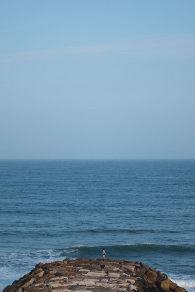A person stands at the end of a rocky pier, facing the vast blue ocean under a clear skyโa tranquil scene reminiscent of the Best Beaches in Ericeira, where waves gently approach the shore and the horizon stretches endlessly.