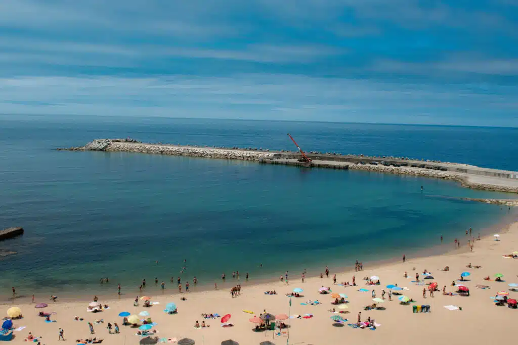 A sandy beach with colorful umbrellas and sunbathers beside calm blue-green water; people are swimming and relaxing. A stone pier with a crane extends into the sea under a partly cloudy sky—one of the Best Beaches in Ericeira.