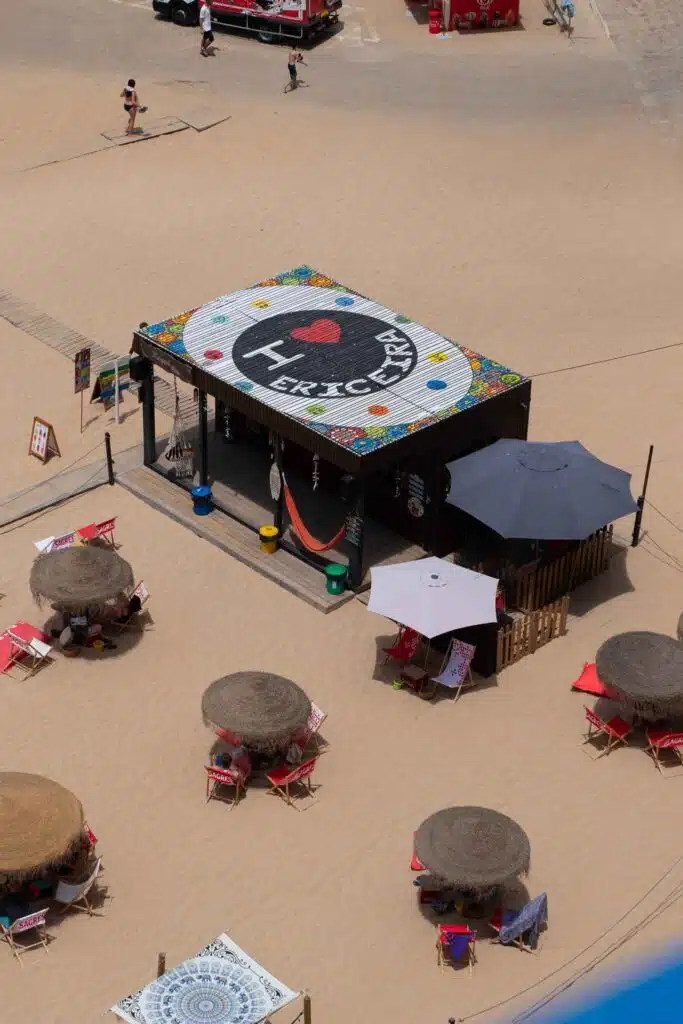 An aerial view of a beach bar with a large roof sign that reads "I ♥ Ericeira." Surrounded by straw umbrellas, lounge chairs, and a sandy beach, it's the perfect spot to experience one of the Best Beaches in Ericeira.