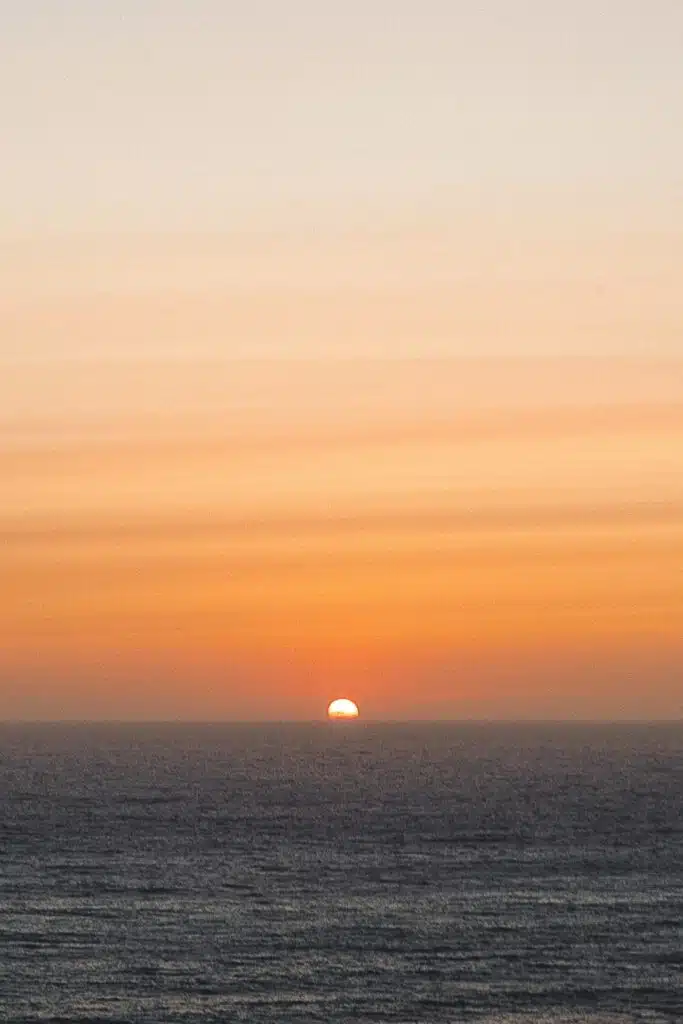 The sun is halfway below the horizon, casting orange and yellow hues across the sky and reflecting on the calm ocean water—an inspiring sunset for any digital nomad in Ericeira.