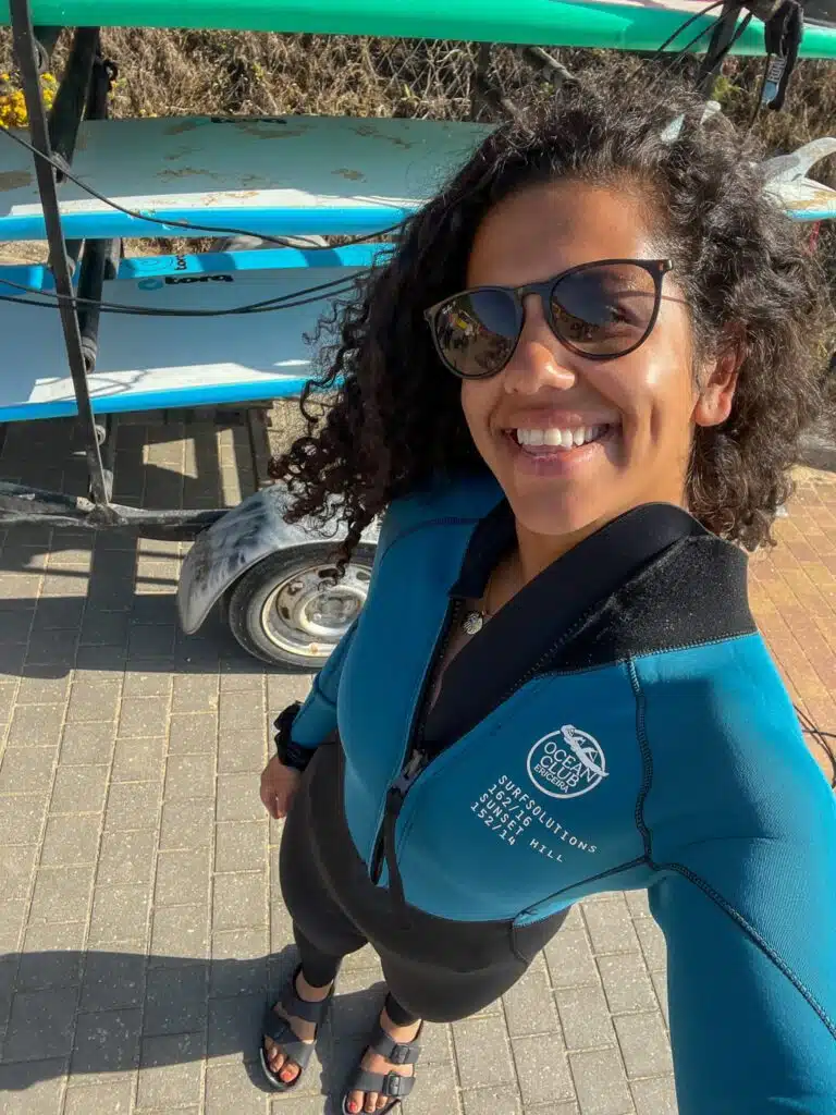 A woman wearing sunglasses and a blue wetsuit smiles for a selfie outdoors, standing near a rack of surfboards on a paved surface. It’s sunny, and this Digital Nomad in Ericeira looks ready for her next water adventure.