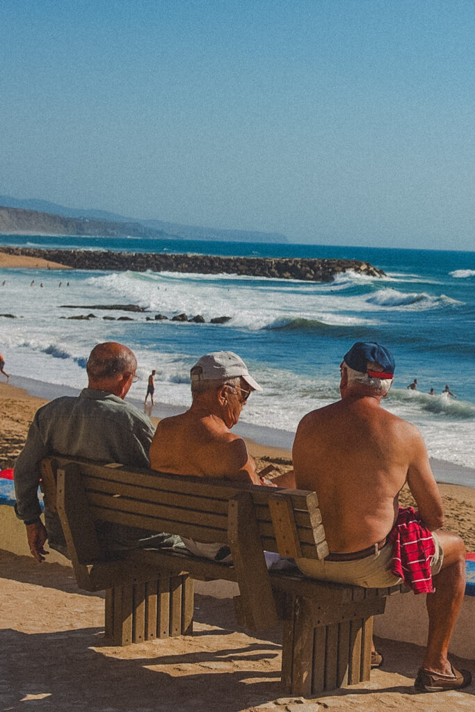 Three older men sit on a wooden bench facing the ocean at one of the Best Beaches in Ericeira, with waves and swimmers in the background on a sunny beach day. Two men are shirtless and one wears a cap.