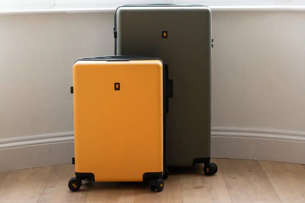 Two Level8 hard-shell suitcases, one yellow and one dark gray, stand side by side on a wooden floor in front of a white wall and window. Both have four wheels and a small square logo in the center.