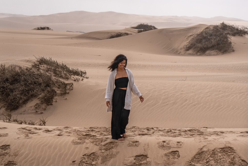 A woman with curly hair, wearing a light shirt over a black top and pants, walks barefoot on sandy dunes with scattered bushes, under a cloudy sky.