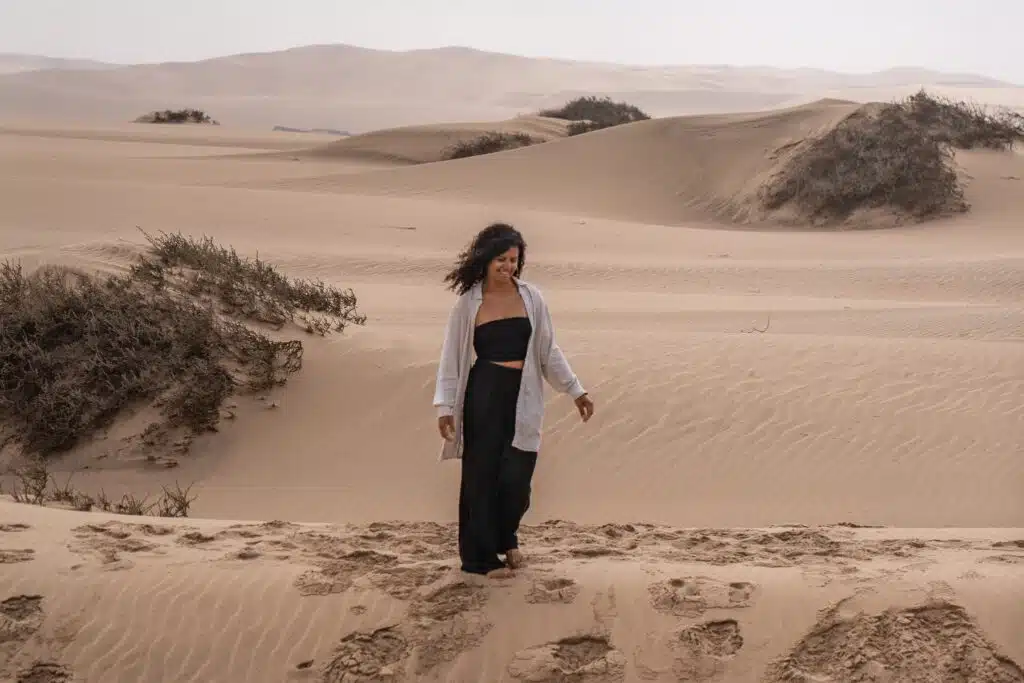 A woman with curly hair, wearing a light shirt over a black top and pants, walks barefoot on sandy dunes with scattered bushes, under a cloudy sky.