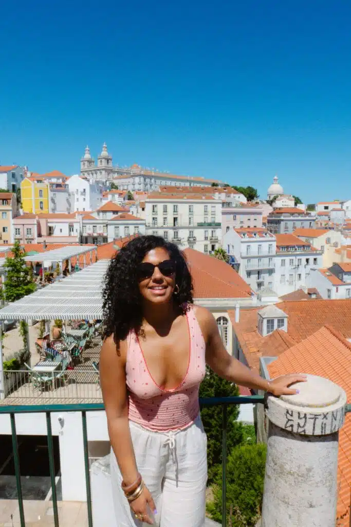 A woman with curly hair and sunglasses, fueled by the courage to travel solo, smiles at a scenic overlook with colorful buildings and terracotta rooftops behind her under a clear blue sky.