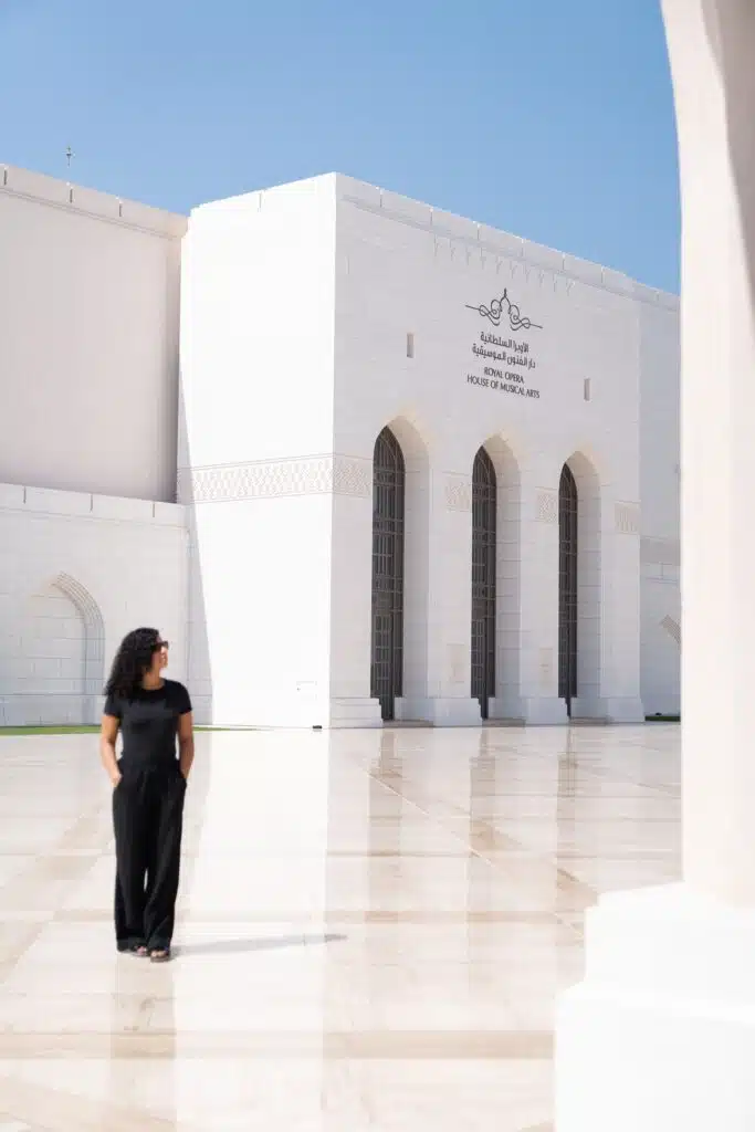 A woman in black clothing, with the courage to travel solo, stands on a polished courtyard in front of the white marble Royal Opera House Muscat, Oman, framed by tall arched doors and a clear blue sky.