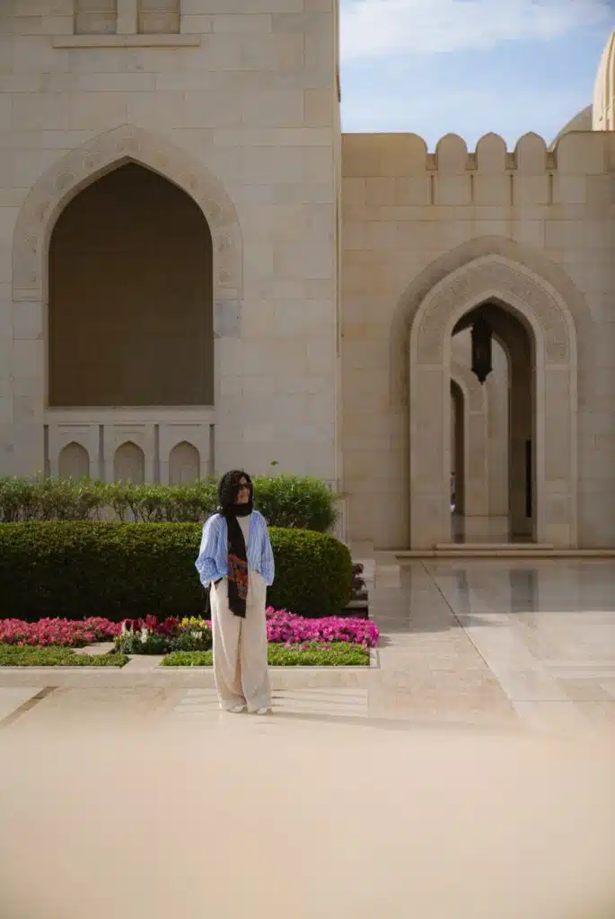 A person with the courage to travel solo, wearing a blue shirt and white pants, stands in front of a beige stone building with arched doorways and blooming flowers under a partly cloudy sky.