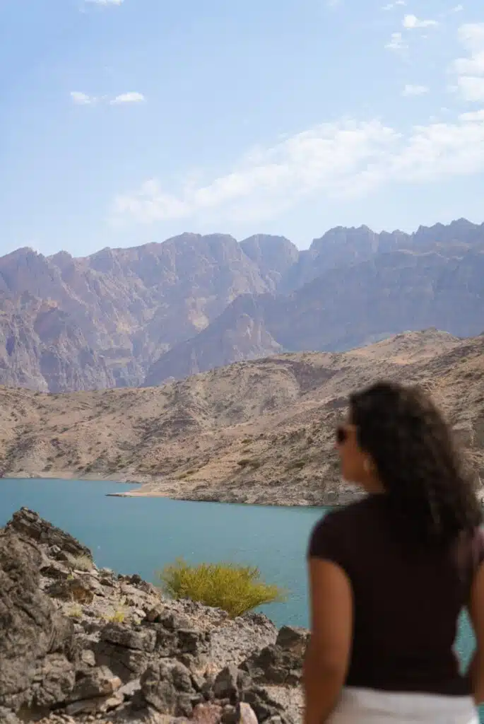 A woman with curly hair stands in the foreground, embodying the courage to travel solo as she gazes at a turquoise lake surrounded by rocky, brown mountains under a partly cloudy sky.