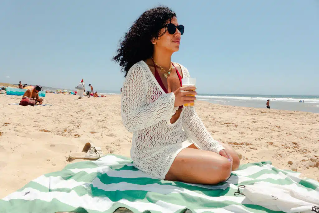 A woman with the courage to travel solo, wearing sunglasses and a white cover-up, sits on a striped towel at the sandy beach, holding a drink and looking to the side as people, umbrellas, and the ocean fill the background.
