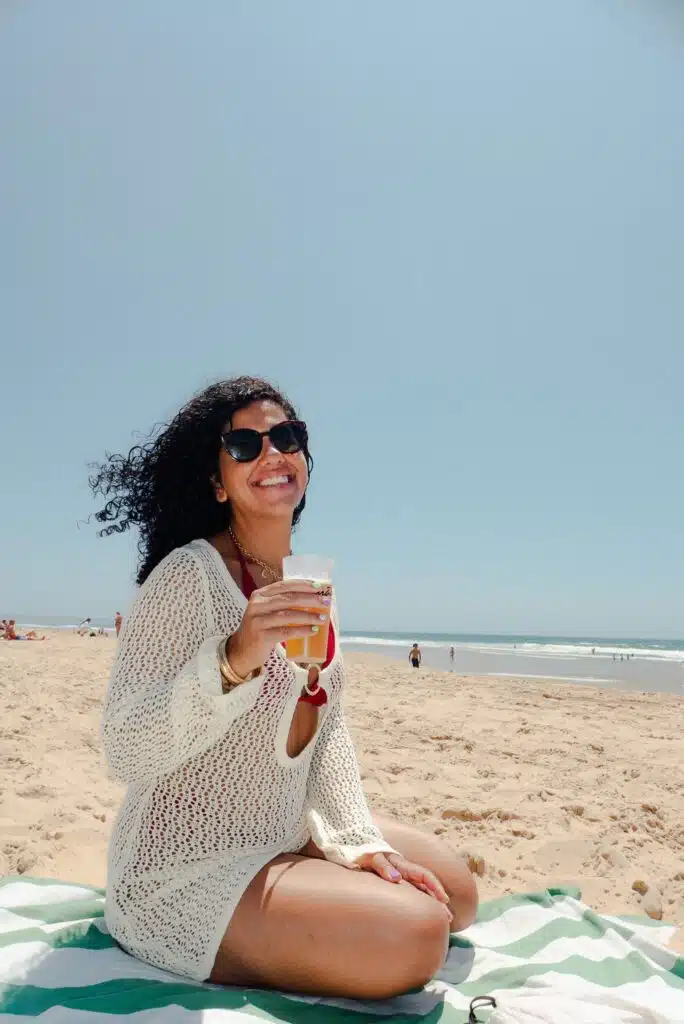 A woman with curly hair and sunglasses sits on a beach towel, smiling and holding a drink. Radiating the courage to travel solo, she wears a white crochet cover-up over a red swimsuit. The sandy beach and blue sky are in the background.