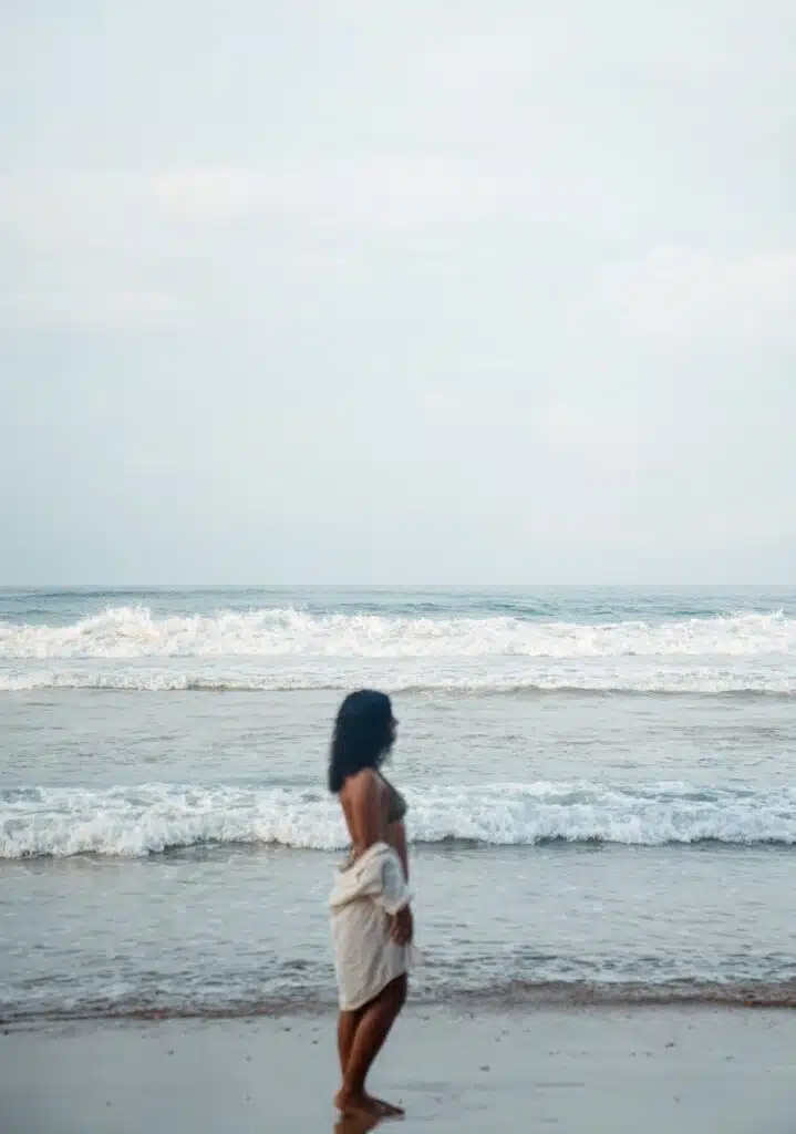 A person wrapped in a towel stands on a sandy beach facing the ocean, with gentle waves and a cloudy sky in the background, embodying the courage to travel solo.