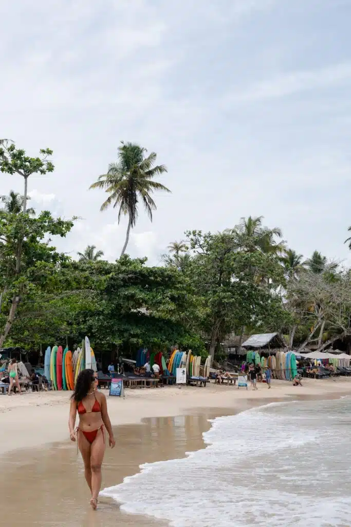 A woman in a red bikini, with the courage to travel solo, walks along a sandy beach near the water. Surfboards, palm trees, and people relaxing under canopies fill the vibrant, partly cloudy backdrop.