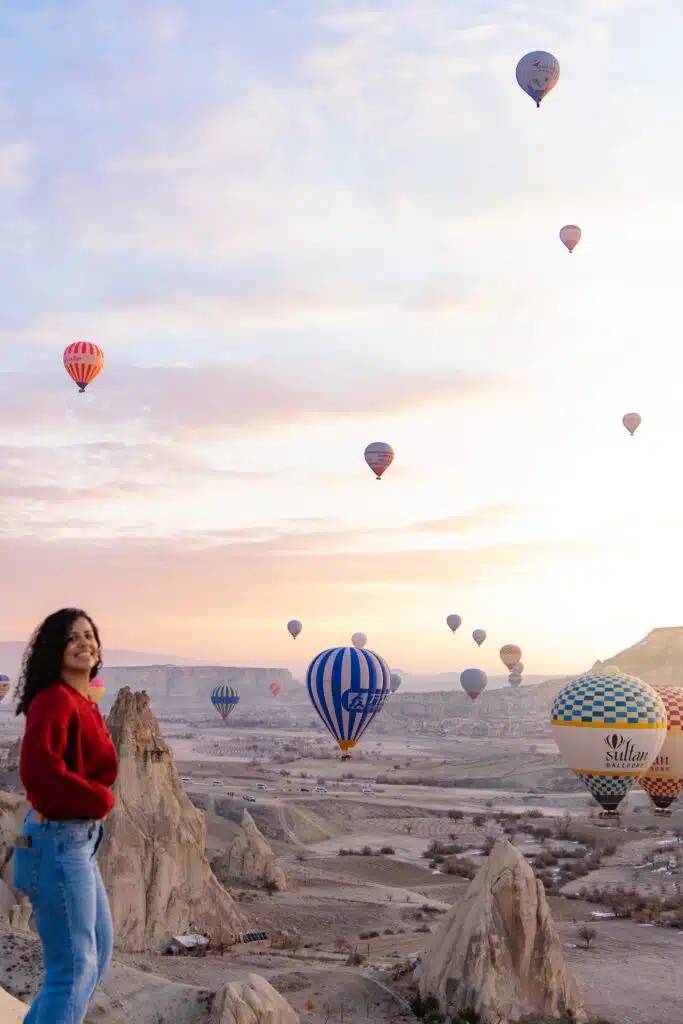 A woman in a red sweater stands on a rocky ledge, smiling with the courage to travel solo, as colorful hot air balloons float over a scenic valley at sunrise. The pastel sky and rugged rock formations create a breathtaking backdrop.