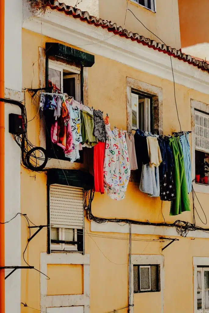 A row of colorful clothes hangs on a line outside the windows of a yellow building in Lisbon, drying in the sunlight—an everyday scene you might spot during 3 days in Lisbon, with electrical wires running along the facade.