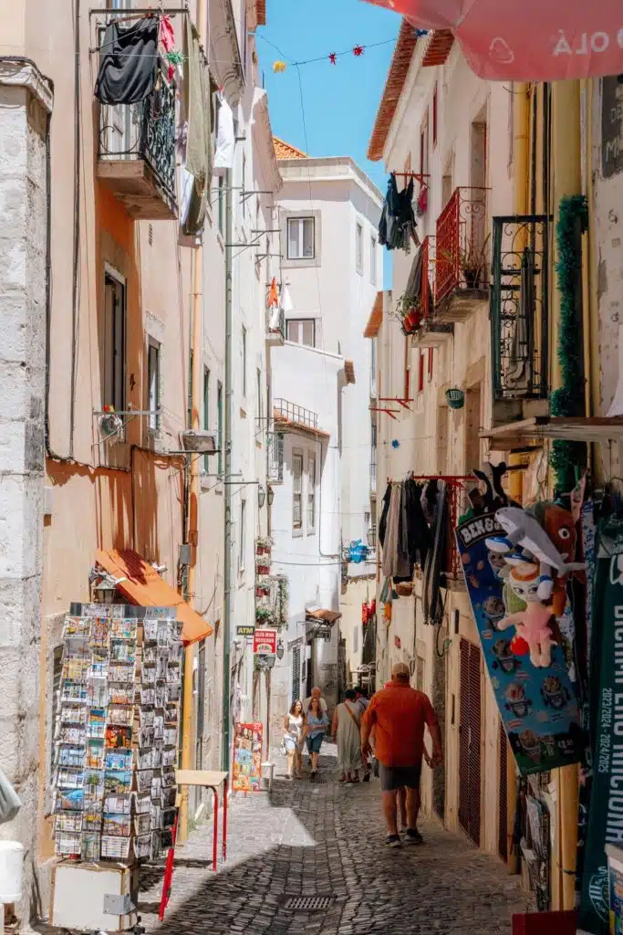 A narrow, sunlit European street in Lisbon is lined with colorful buildings, hanging laundry, balconies, and souvenir stands. People stroll the cobblestone path, savoring the lively atmosphere—perfect for anyone spending 3 days in Lisbon.
