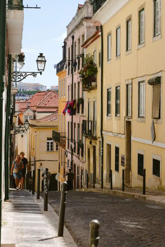 A narrow, cobblestone street in a European city—perfect for exploring during 3 days in Lisbon—with pastel yellow buildings, red tiled roofs, balconies bursting with flowers, and people strolling uphill on a sunny day.