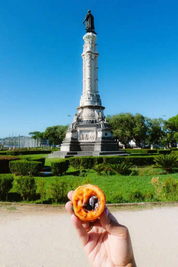 A hand holds up a pastel de nata (Portuguese custard tart) in front of a tall white monument with a statue on top—an iconic moment from 3 days in Lisbon, set in a green park under a clear blue sky.