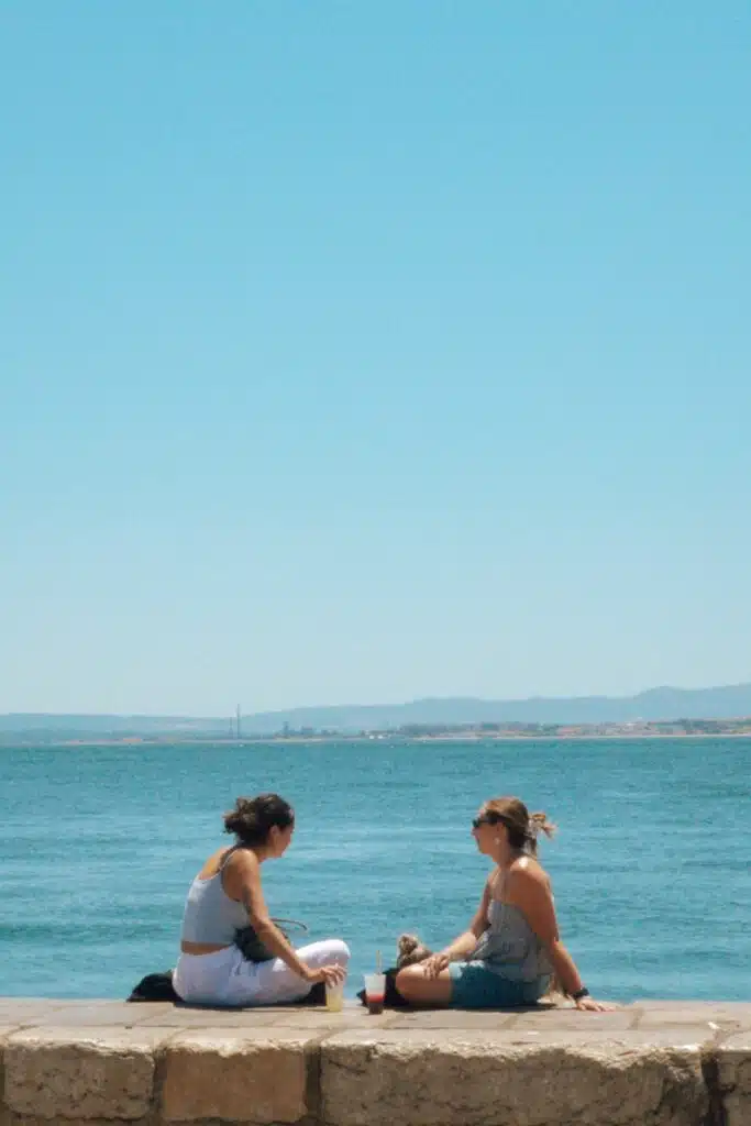 Two women sit on a stone ledge by the water, relaxing and sharing stories about their 3 days in Lisbon. Drinks rest between them as they enjoy the clear blue sky and distant mountain views.